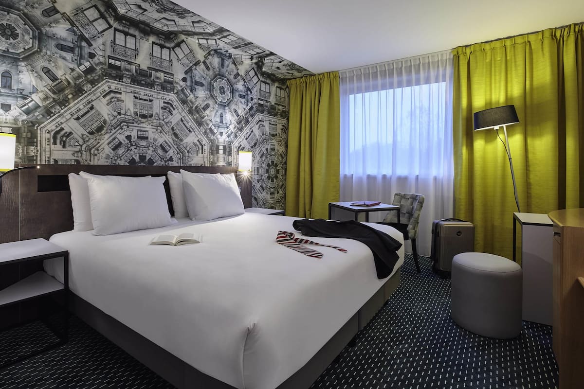 Hotellihuone, jossa on suuri sänky, työpöytä, tuoli, lamppu ja ikkunasta avautuva näkymä ulos, ikkunassa on verhot.