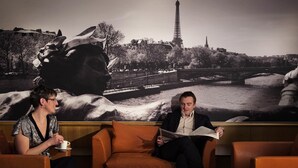 Deux personnes assises sur un canapé, une table devant elles, une grande fresque murale en arrière-plan et une vue sur la Tour Eiffel.