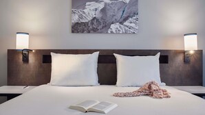 Une chambre d’hôtel avec un grand lit, deux oreillers, une couverture et un livre ouvert. Il y a deux lampes fixées au mur et un tableau encadré au-dessus du lit.