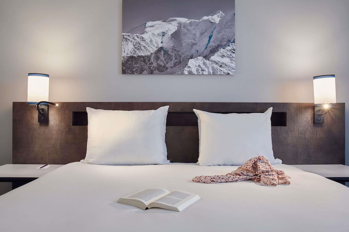 Habitación de hotel con una cama grande, dos almohadas, una colcha y un libro abierto. Hay dos lámparas de pared y un cuadro enmarcado sobre la cama.
