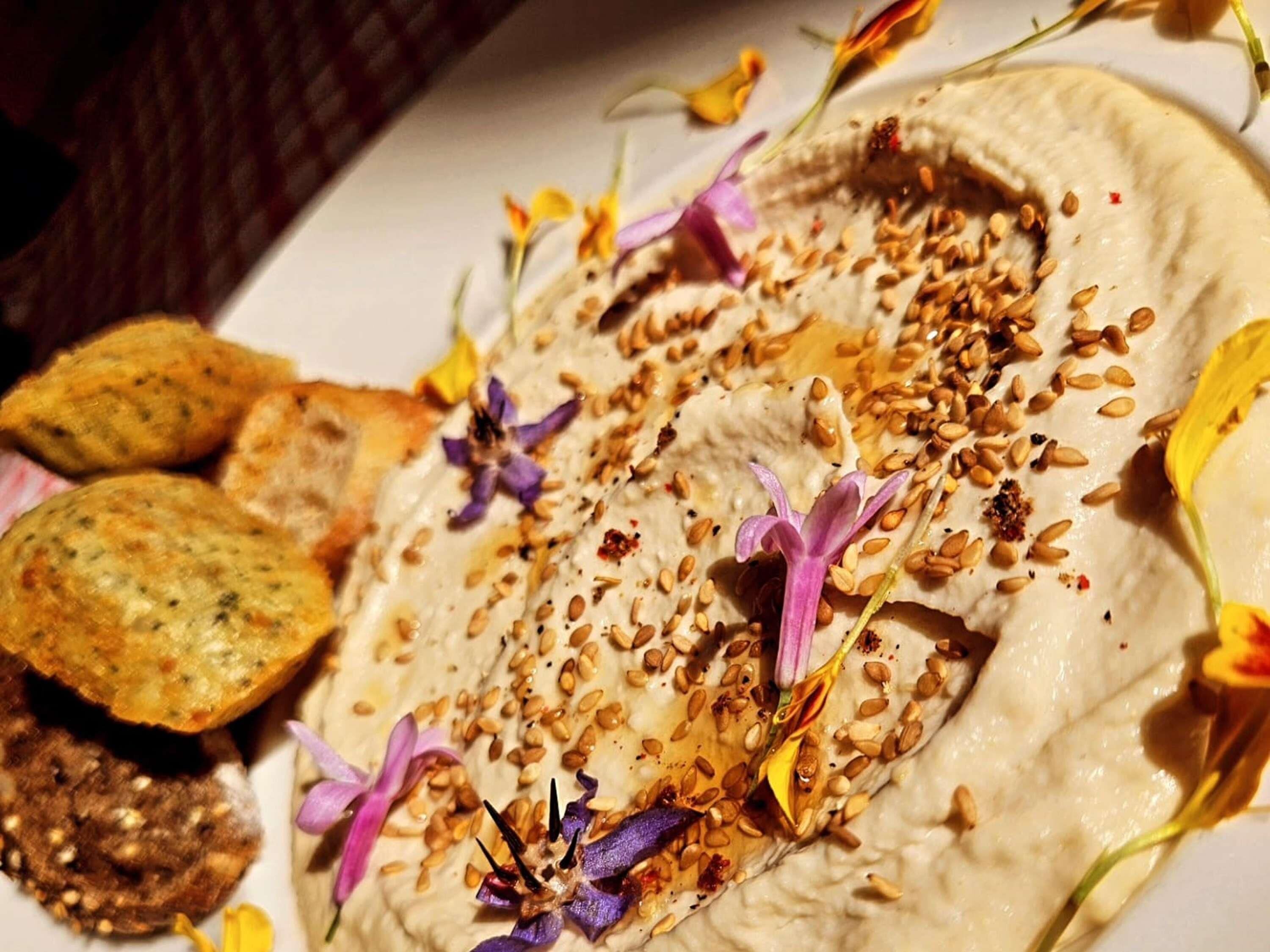 A plate of hummus with sesame seeds and edible flowers, accompanied by pita bread and a side of olives.