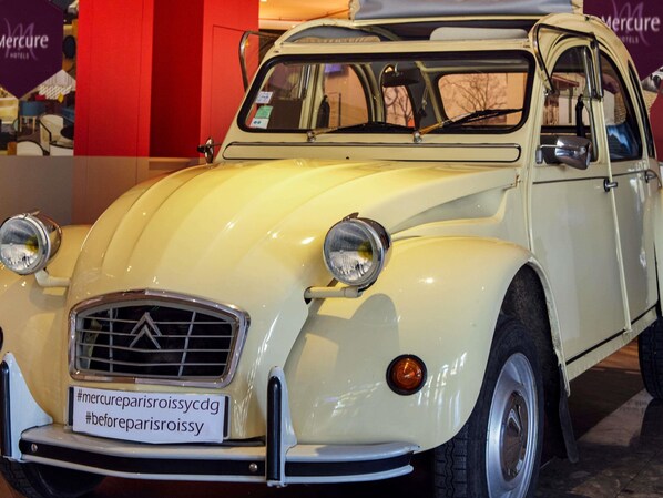 Un coche antiguo con un cartel que dice '#mercuryparisroissycdg #beforeparisroissy' frente a un hotel.