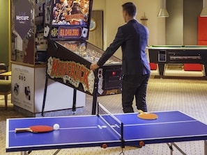 Un hombre jugando ping pong en el vestíbulo de un hotel, con una mesa de billar al fondo.