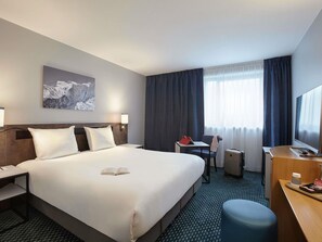 Habitación de hotel con una cama grande, un escritorio con computadora, una silla, un televisor y una ventana con cortinas.