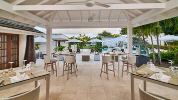 Un espacio de comedor al aire libre con techado, mesas y sillas, una piscina al fondo y sombrillas.