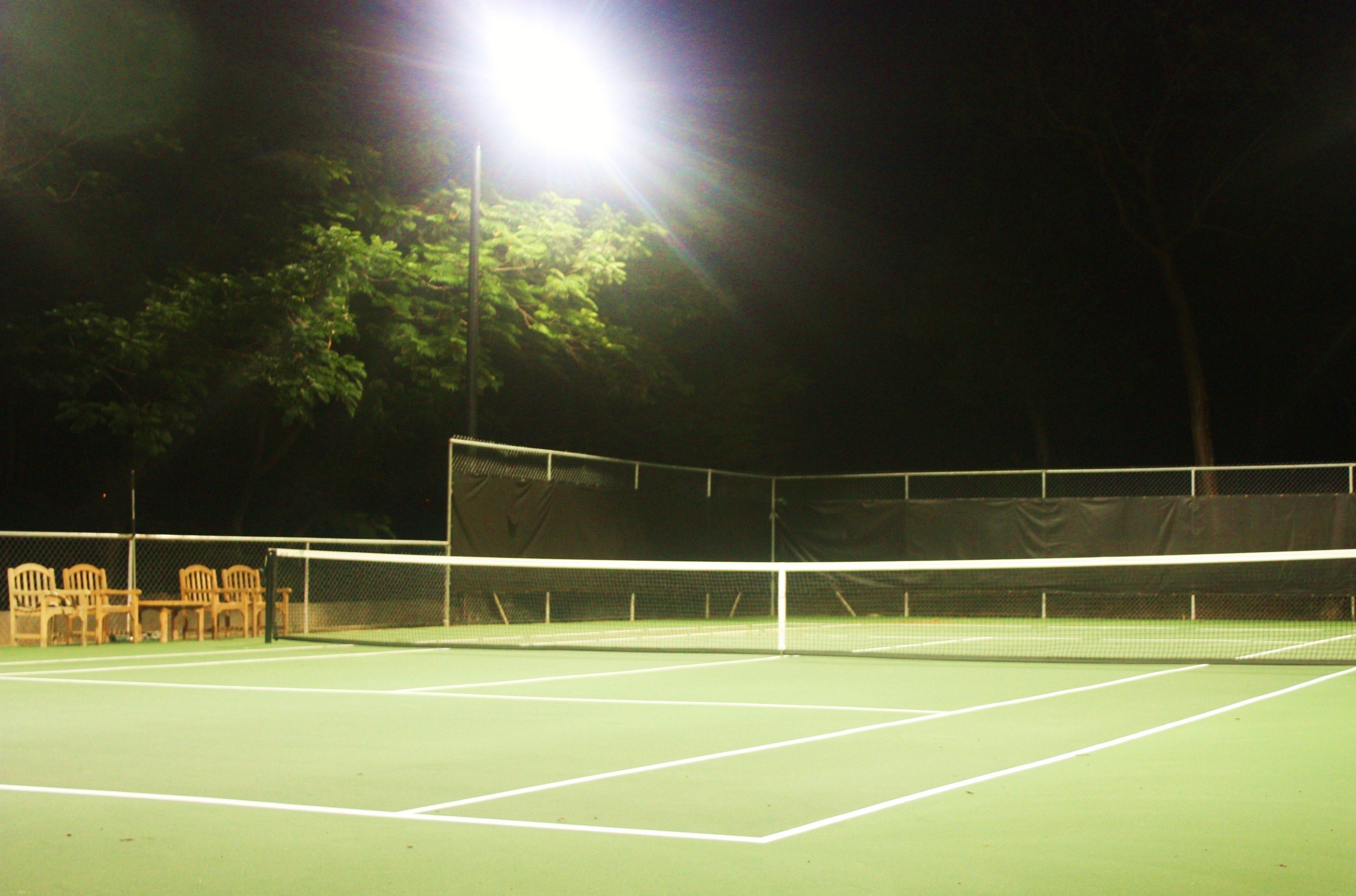 A tennis court at night with bright floodlights, a net, and chairs along the side.