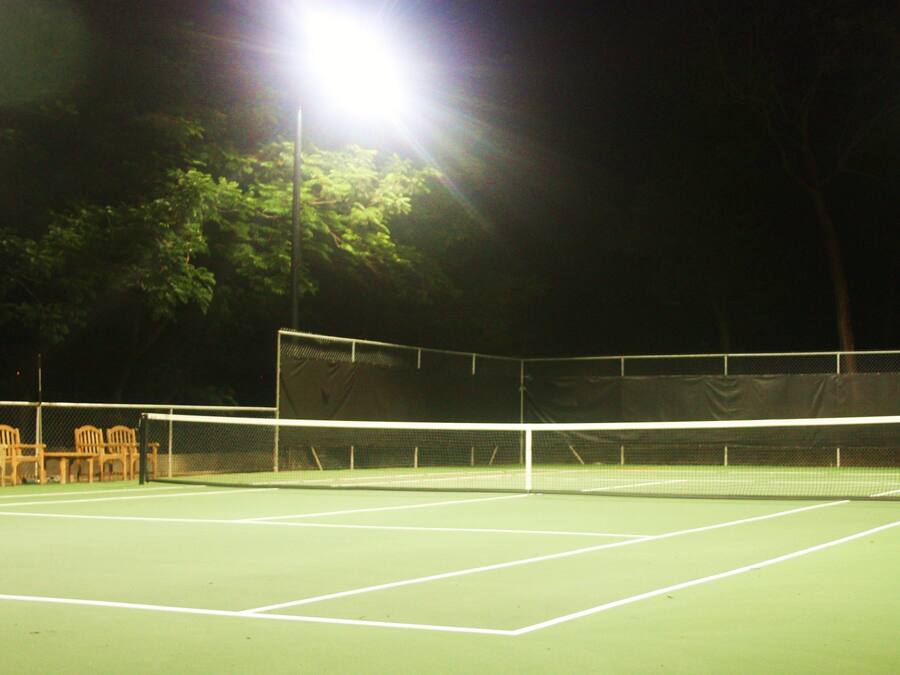 A tennis court at night with bright floodlights, a net, and chairs along the side.