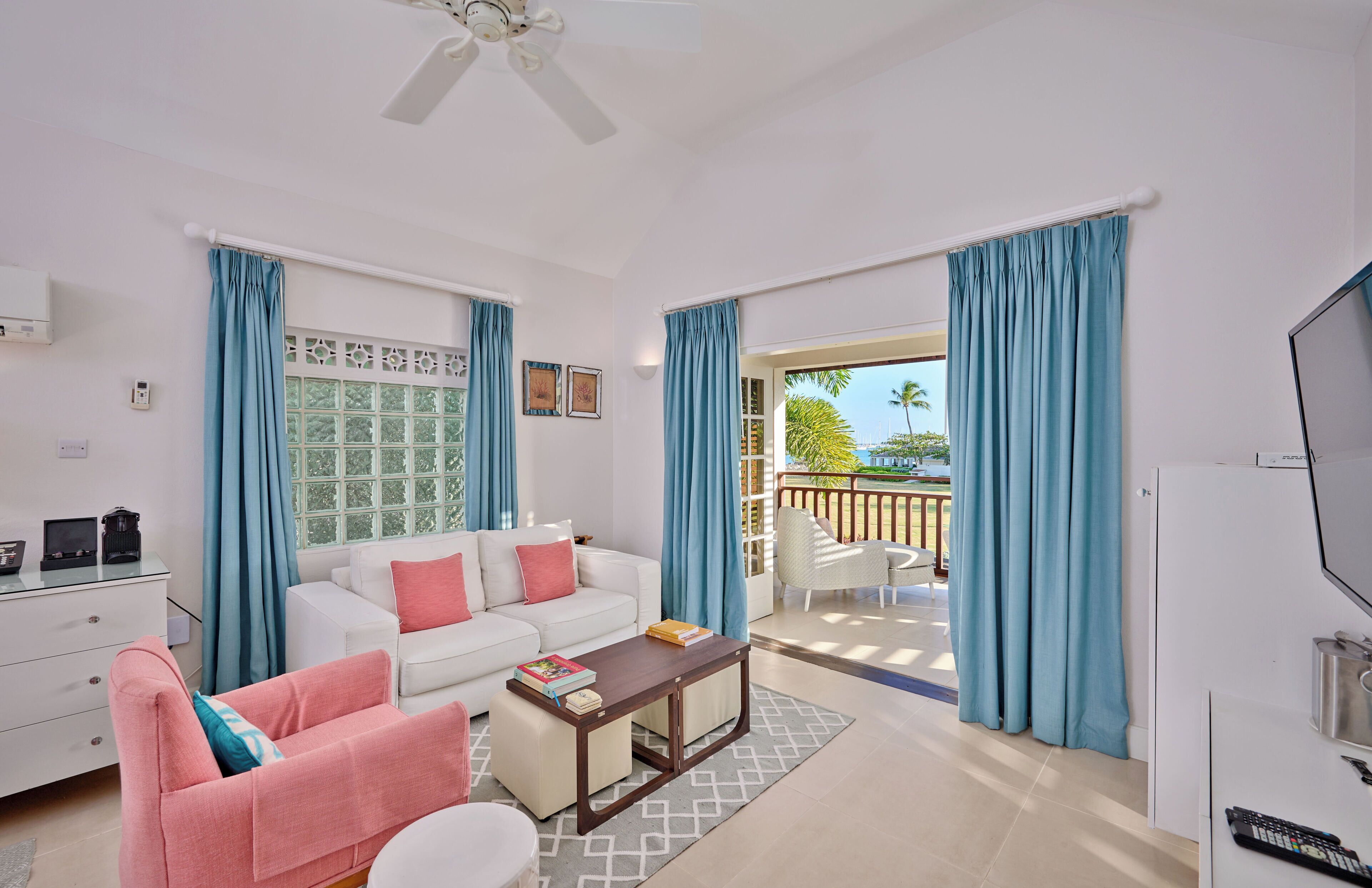 A modern living room with a white sofa, a pink armchair, a coffee table, and a TV. There is a balcony with blue curtains and a view of greenery.