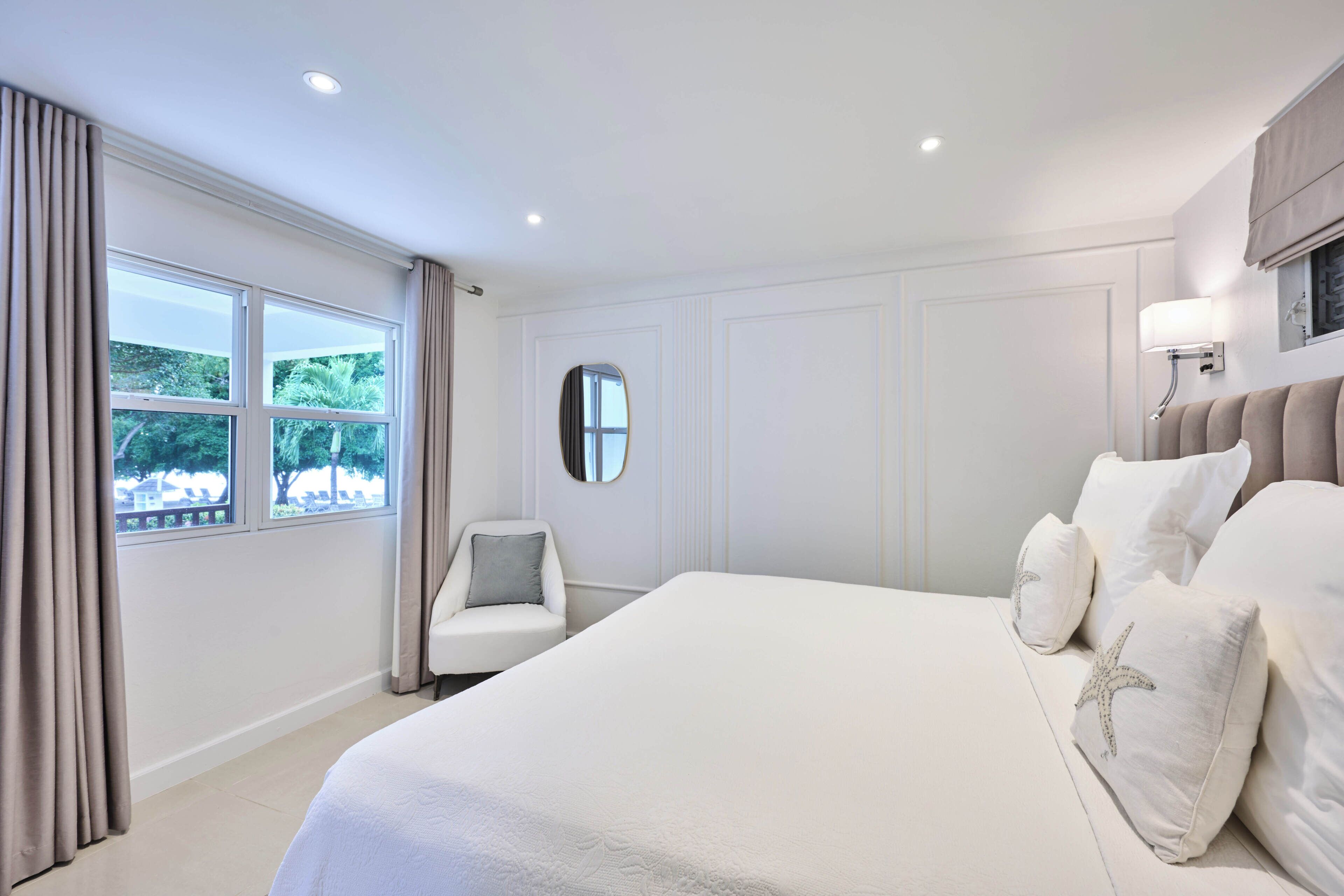 Un dormitorio con una cama grande, una silla, un ventanal con cortinas, un espejo y una lámpara fijada a la pared.