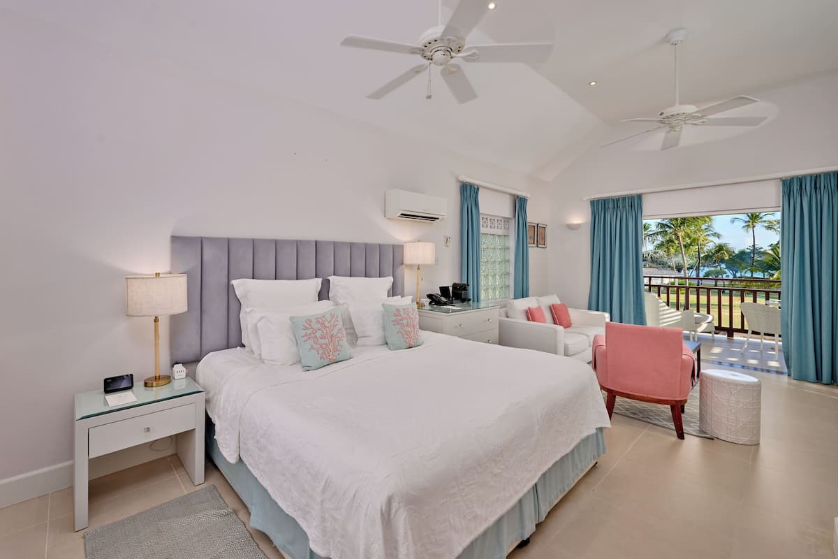 A hotel room with a large bed, two ceiling fans, a desk with a lamp, a chair, a small table, and a view of palm trees through the window.