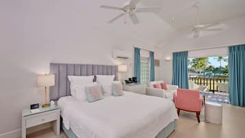 A hotel room with a large bed, two ceiling fans, a desk with a lamp, a chair, a small table, and a view of palm trees through the window.