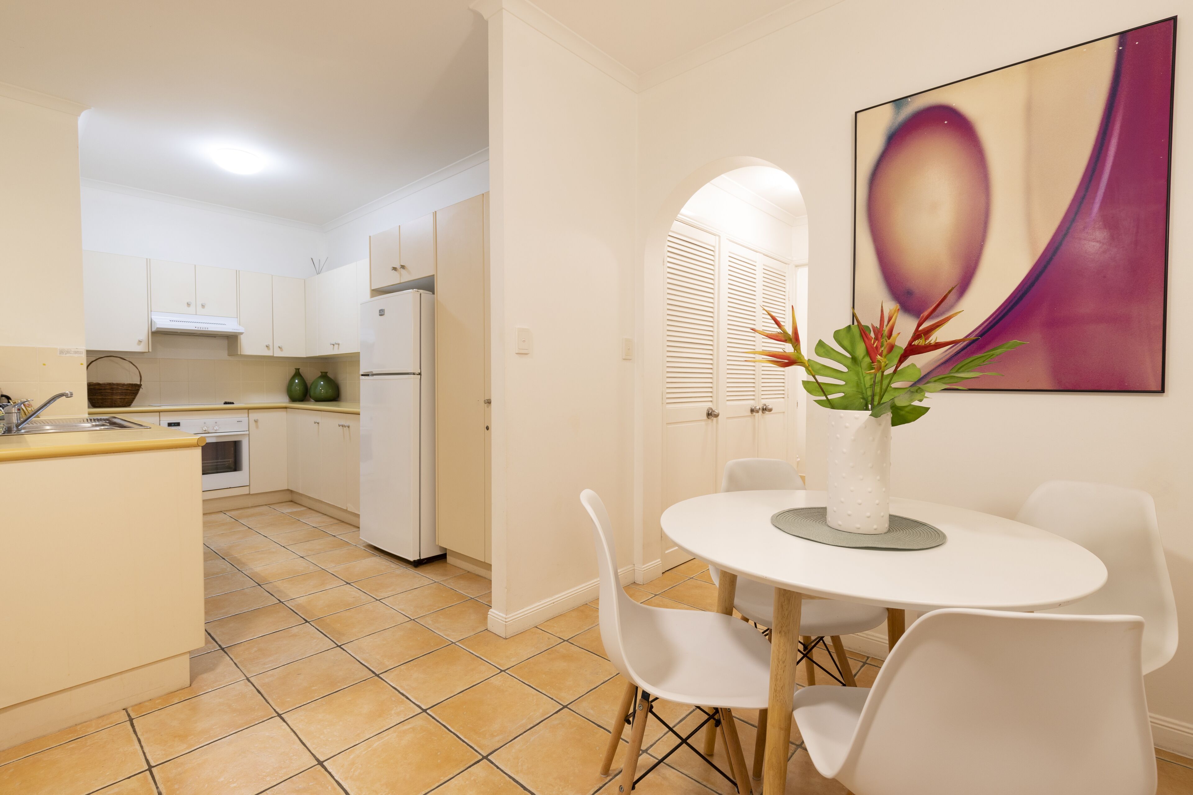 Une cuisine moderne avec des meubles blancs, une table à manger avec des chaises et un grand tableau abstrait accroché au mur.