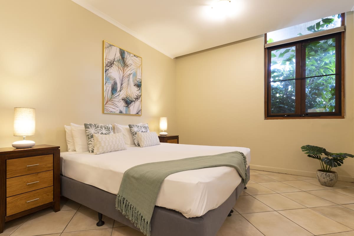 A bedroom with a bed, nightstands, a lamp, a painting, a window, and a potted plant.