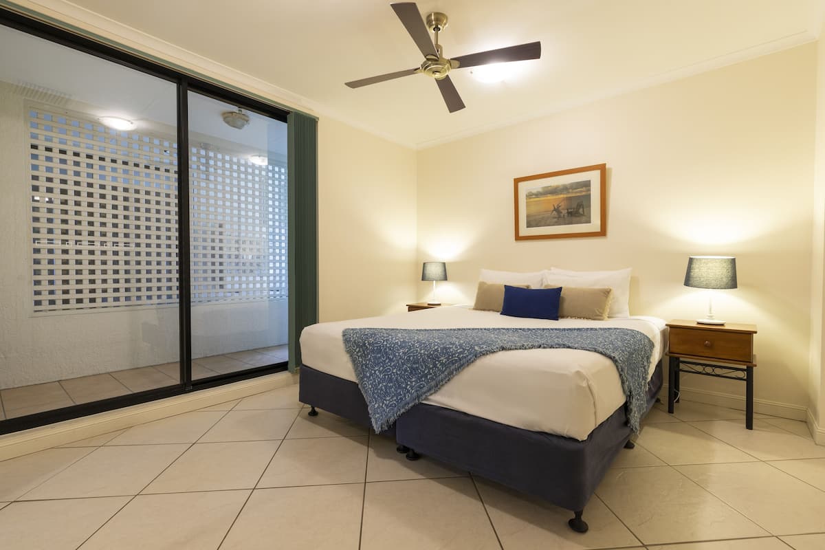 A bedroom with a bed, two bedside tables with lamps, a ceiling fan, a window with a view, and a door leading to a balcony.