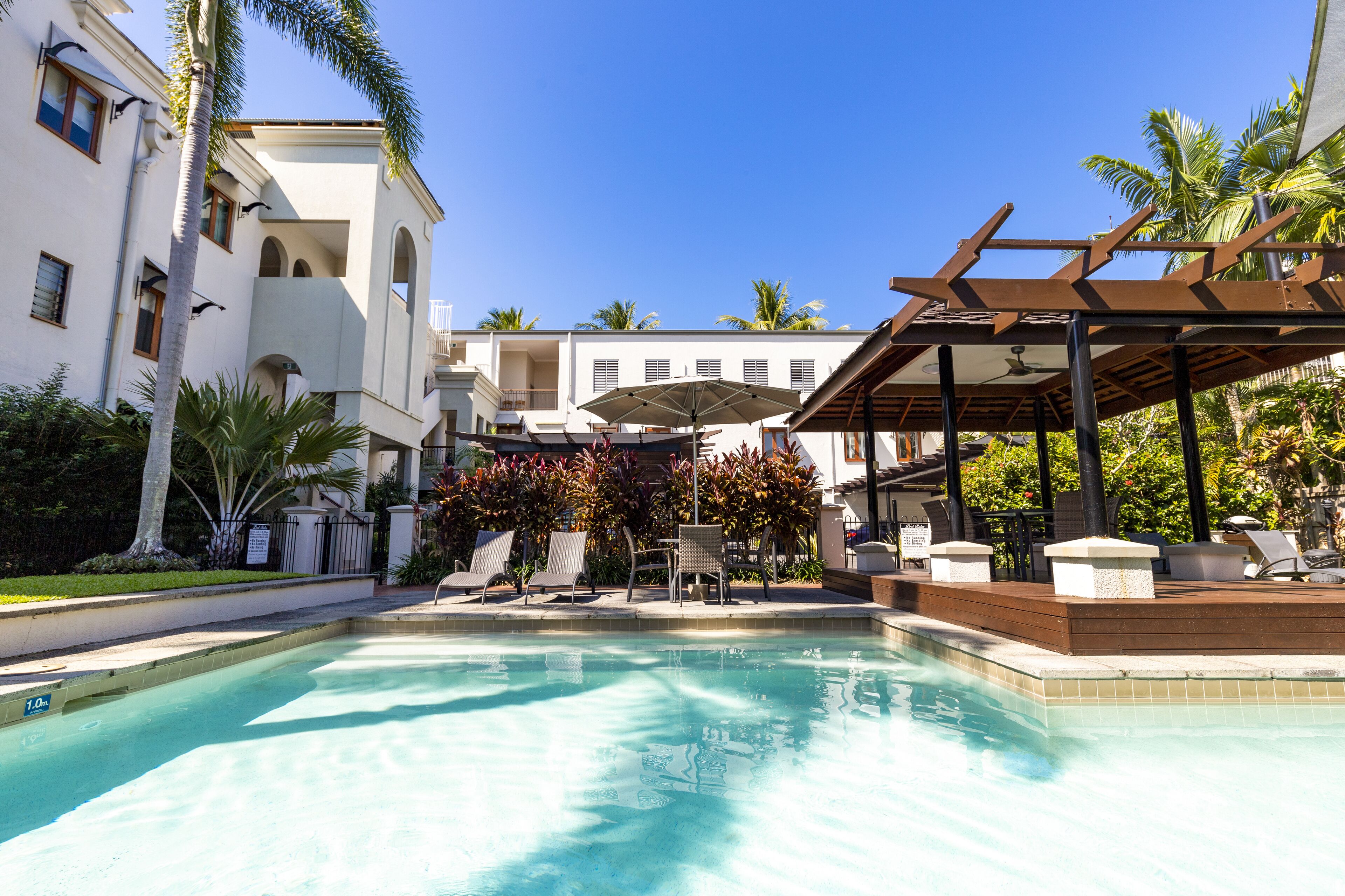 Une piscine entourée de palmiers et d’un bâtiment doté de balcons, avec des chaises longues et des parasols à proximité.