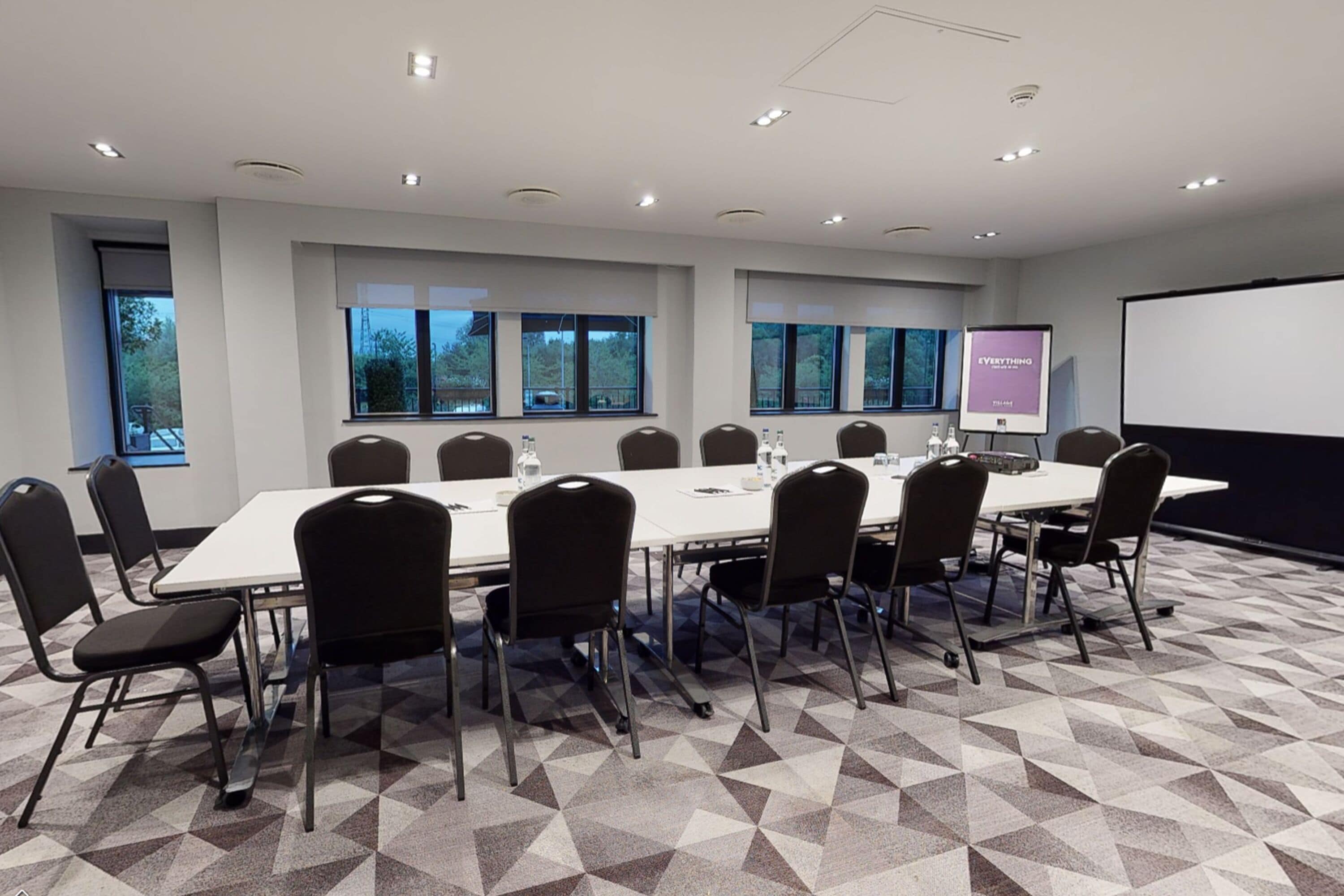 Ein Konferenzraum mit einem langen Tisch, Stühlen, einer Projektionsleinwand und Fenstern mit Blick auf Bäume.