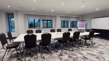 Ein Konferenzraum mit einem langen Tisch, Stühlen, einer Projektionsleinwand und Fenstern mit Blick auf Bäume.