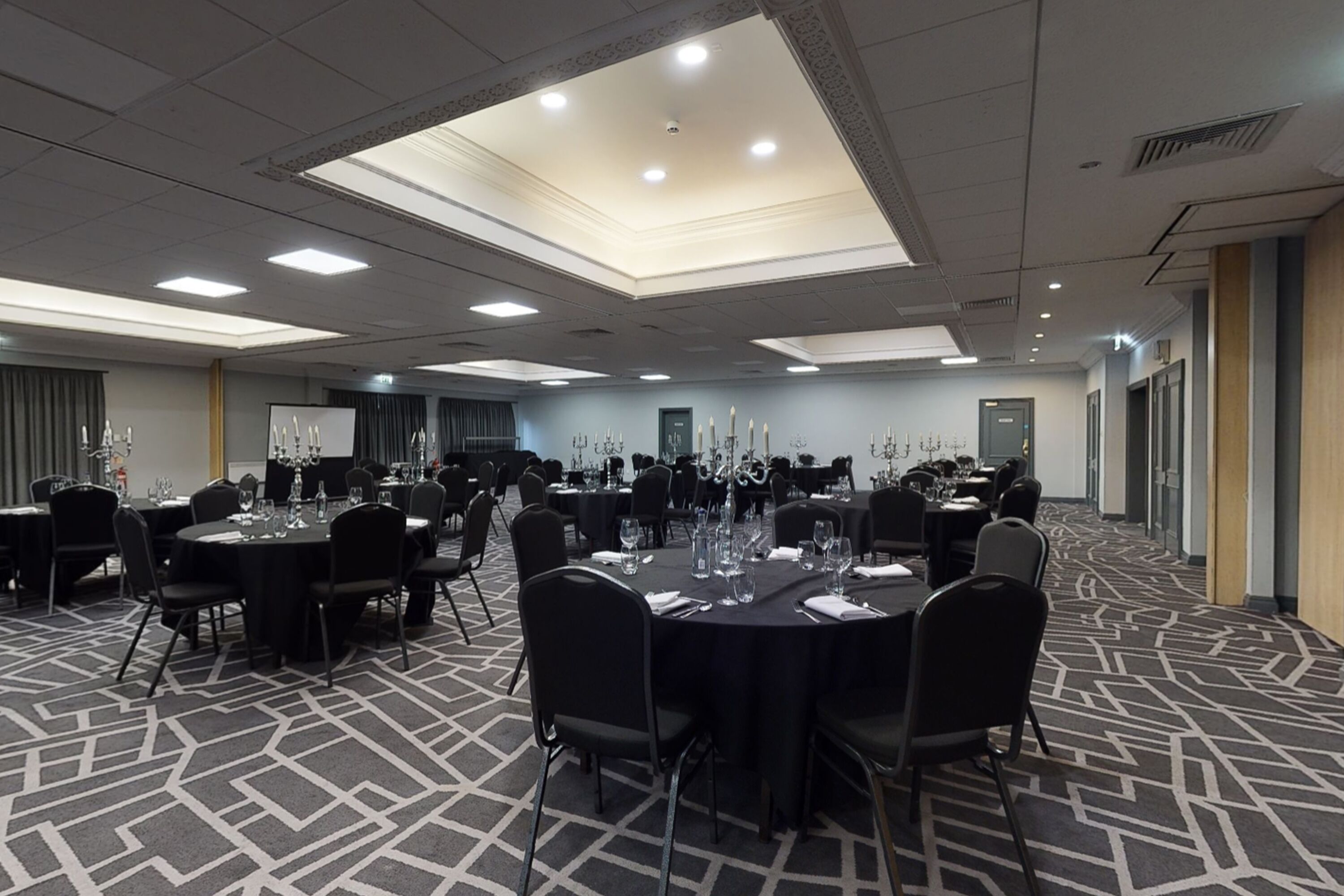 A conference room with multiple round tables, each set with chairs, water bottles, and glassware. The room has a patterned carpet and recessed ceiling lighting.