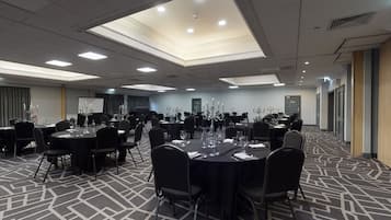 A conference room with multiple round tables, each set with chairs, water bottles, and glassware. The room has a patterned carpet and recessed ceiling lighting.