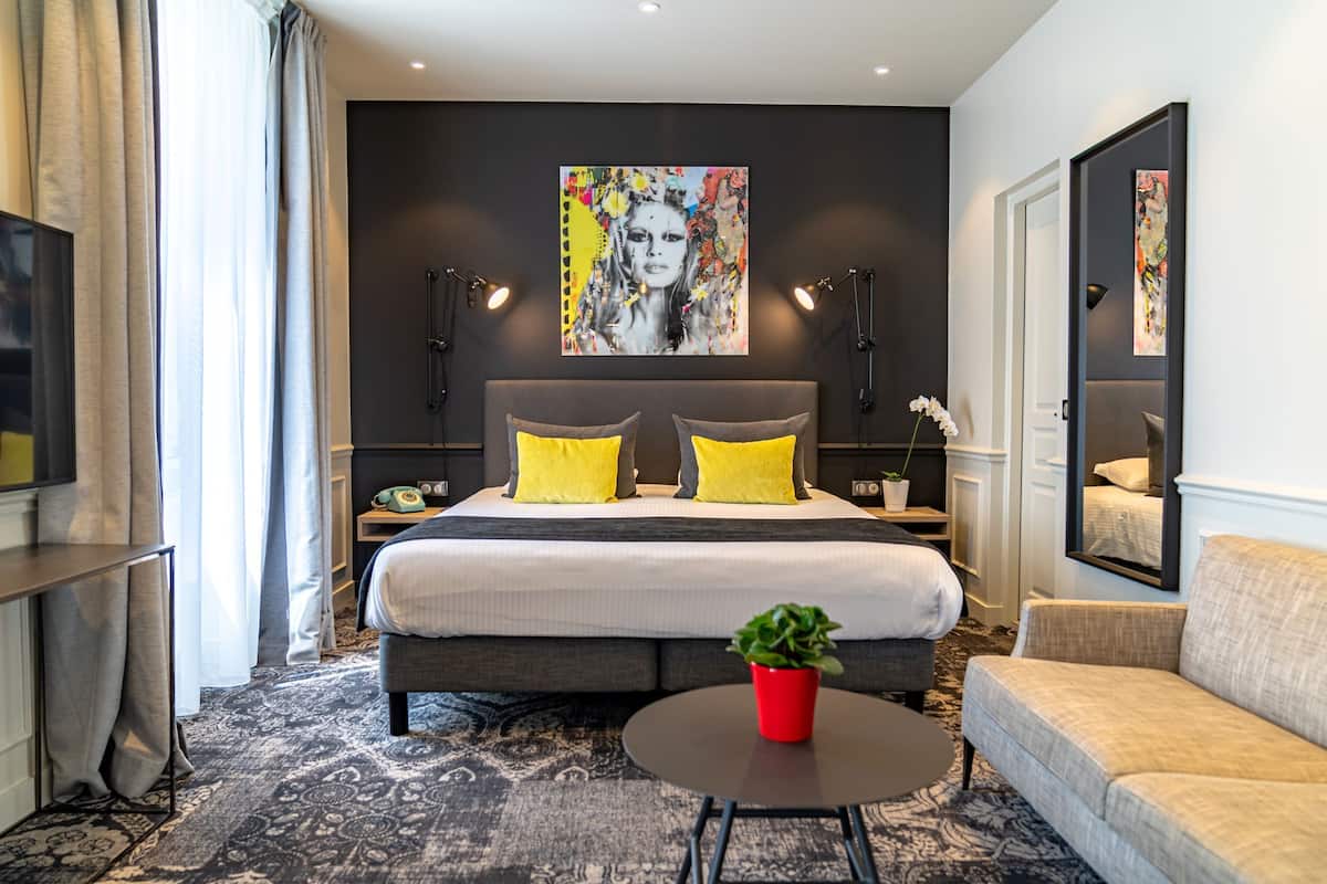 A modern hotel room with a large bed, two yellow pillows, a red plant on a round table, and a large painting on the wall.
