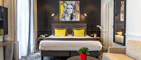 Ein modernes Hotelzimmer mit einem großen Bett, zwei gelben Kissen, einer roten Pflanze auf einem runden Tisch und einem großen Gemälde an der Wand.
