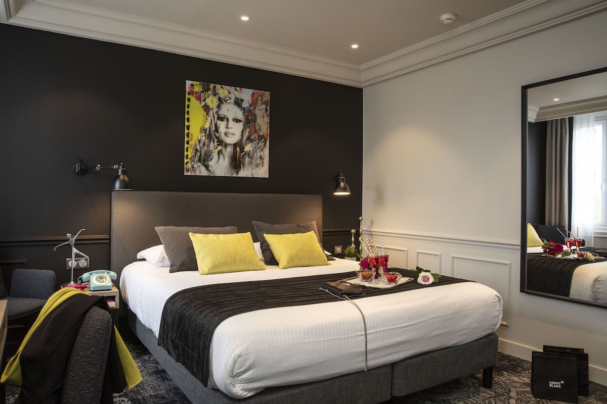 A modern hotel room with a large bed, two bedside lamps, a bedside telephone, a tray table with a drink and flowers, and a large mirror.