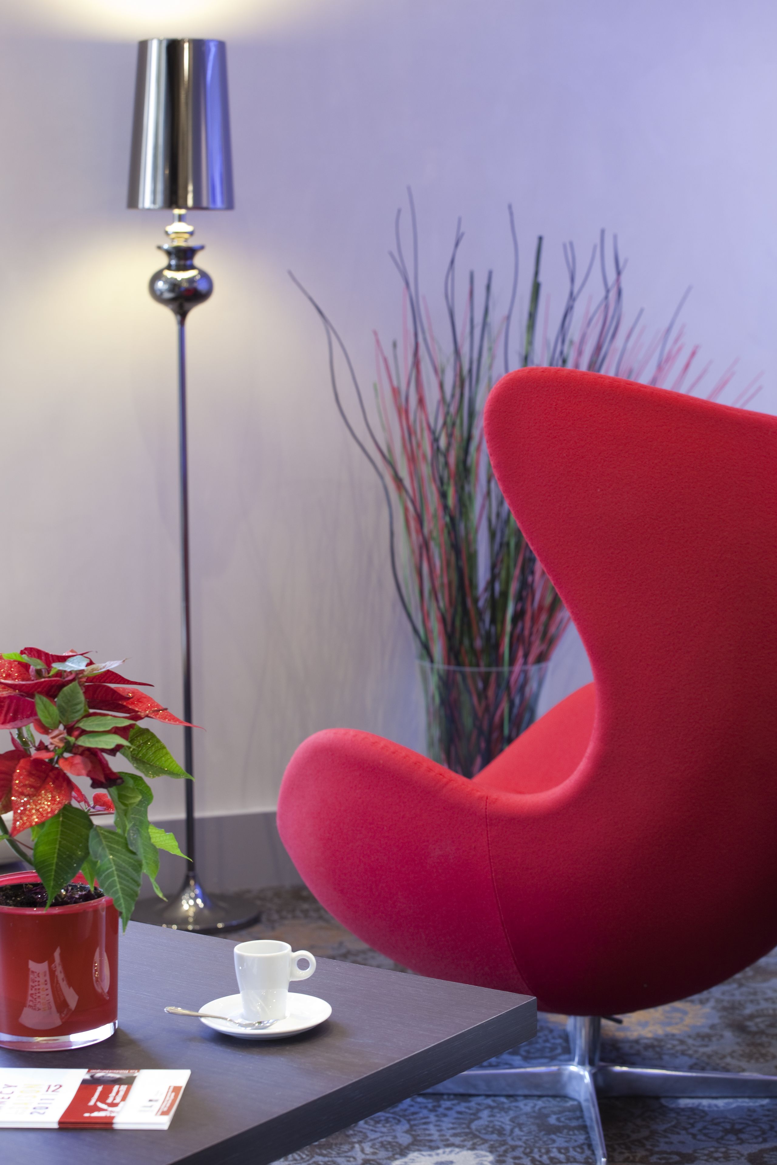 A modern room with a red chair, a small table with a white cup and saucer, a potted plant, and a tall floor lamp.