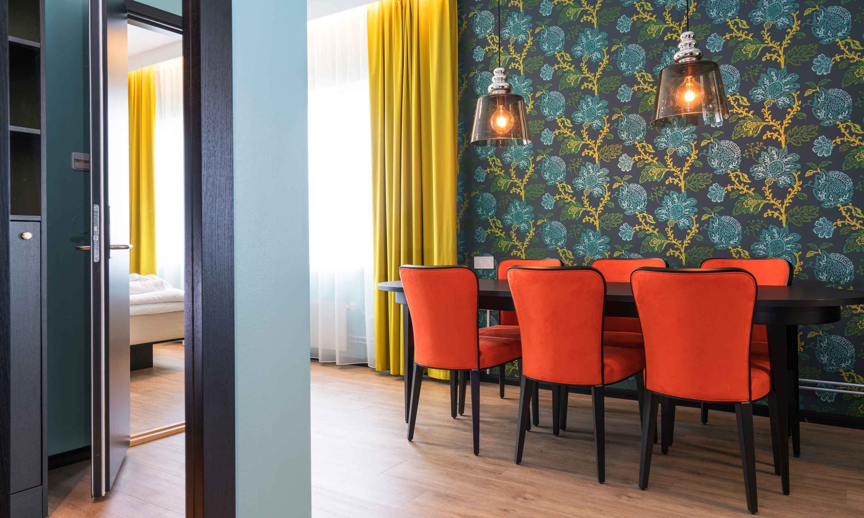 Un comedor moderno con una mesa y sillas, un papel tapiz estampado y vista a una cama a través de una puerta abierta.