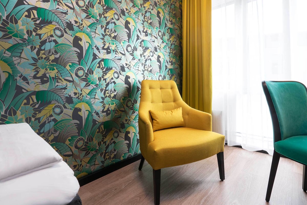 A room with a patterned wallpaper, a yellow armchair, a teal armchair, and a white bed.