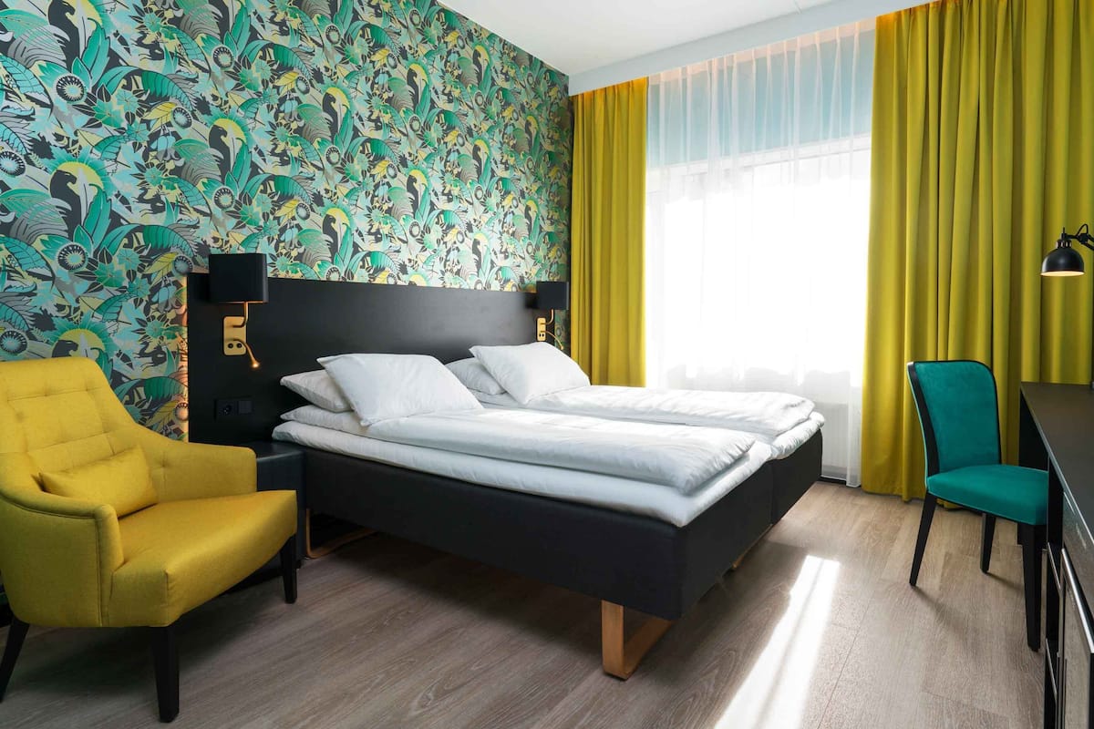 A hotel room with a bed, two chairs, a desk, and a window with yellow curtains.