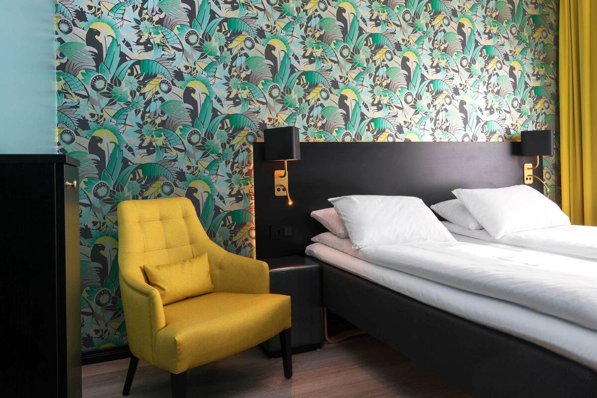 A hotel room with a bed, a yellow armchair, and a wall with a floral pattern.