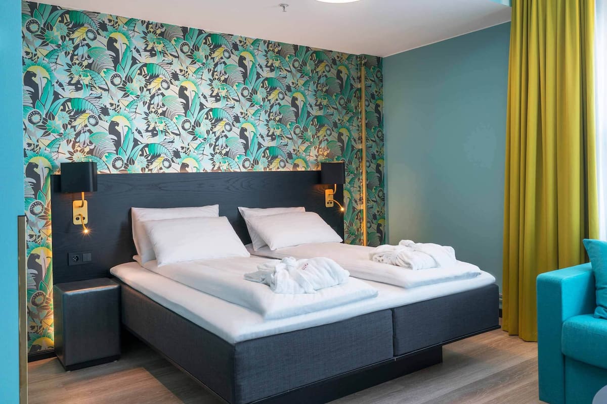 A double bed with white linens, two bedside tables, a wall-mounted lamp, and a teal armchair.