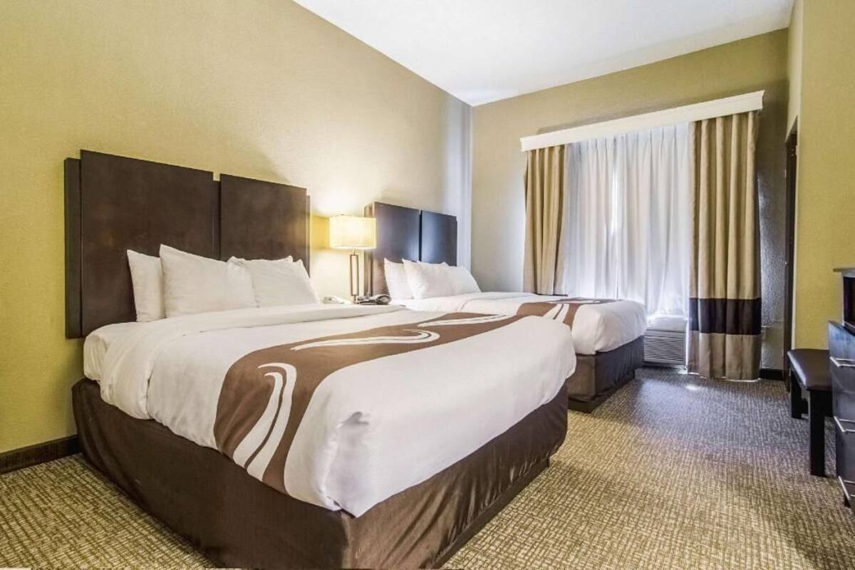 Habitación de hotel con dos camas, un escritorio, una lámpara y una ventana con cortinas.