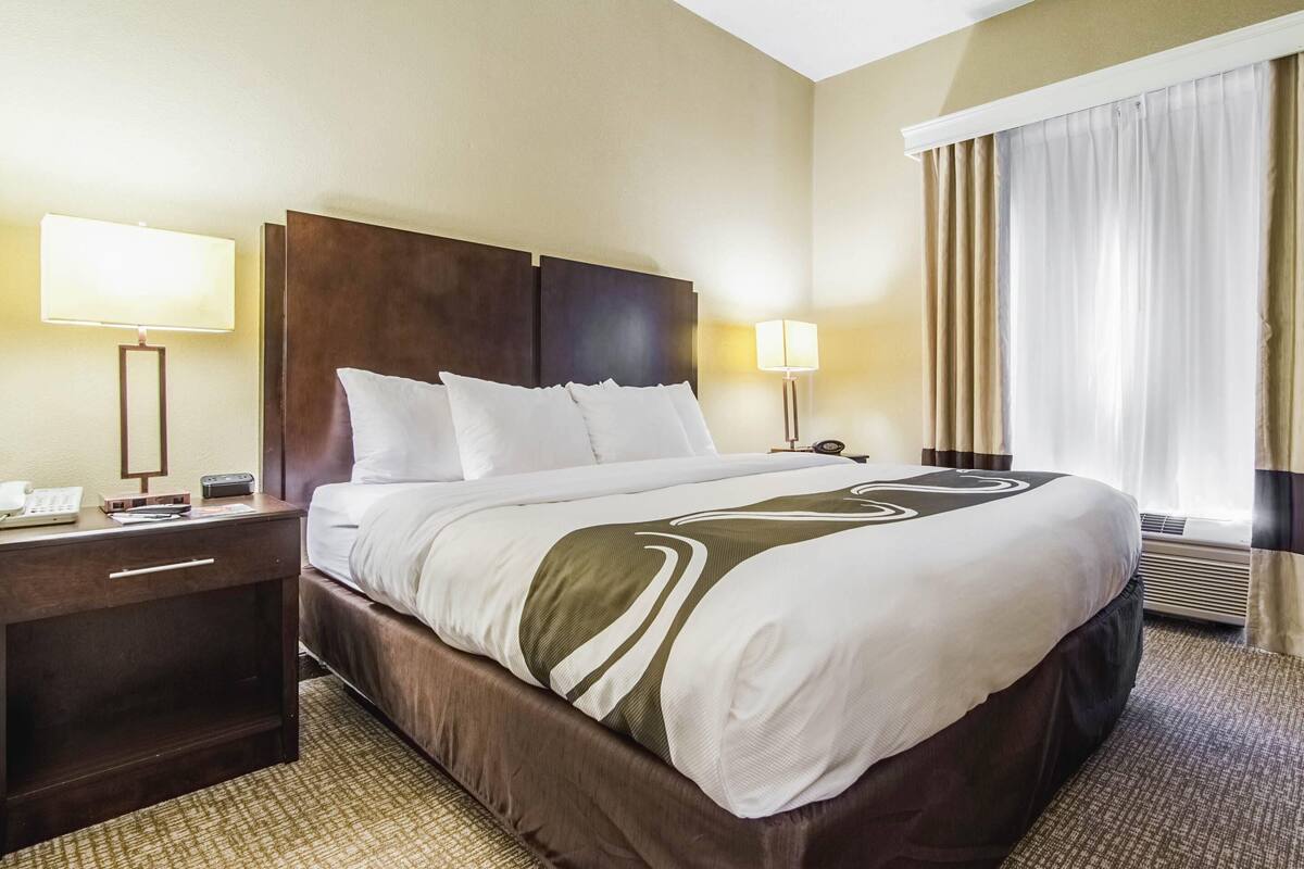 Una habitación de hotel con una cama grande, dos mesitas de noche, una lámpara y una ventana con cortinas.