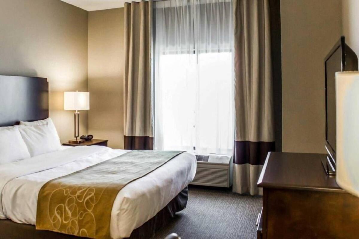Una habitación de hotel con cama, lámpara de noche, escritorio con lámpara, ventana con cortinas y un equipo de aire acondicionado.