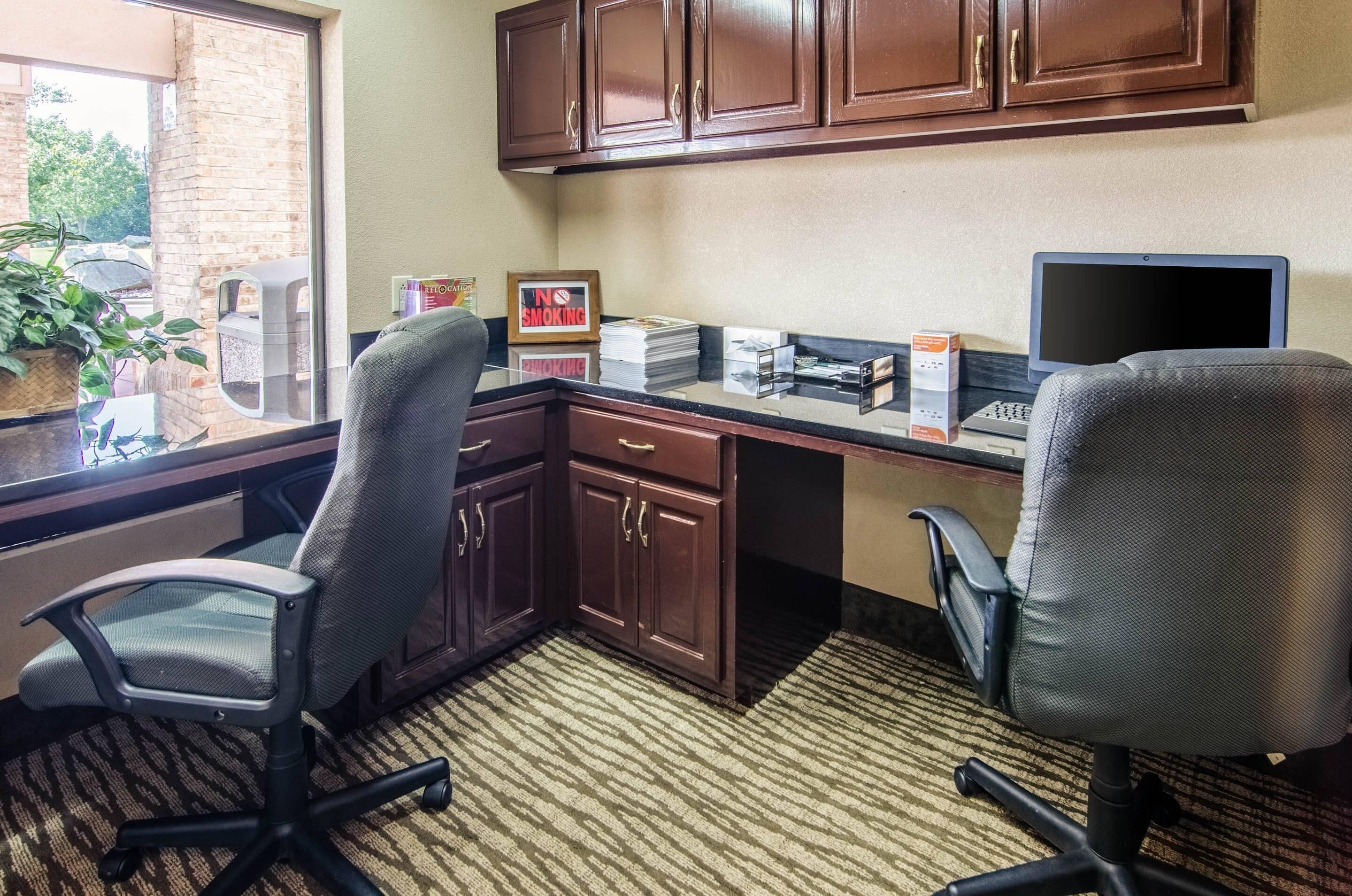 A hotel office with two office chairs, a desk with a laptop, a 'No Smoking' sign, and a window with a view of trees.
