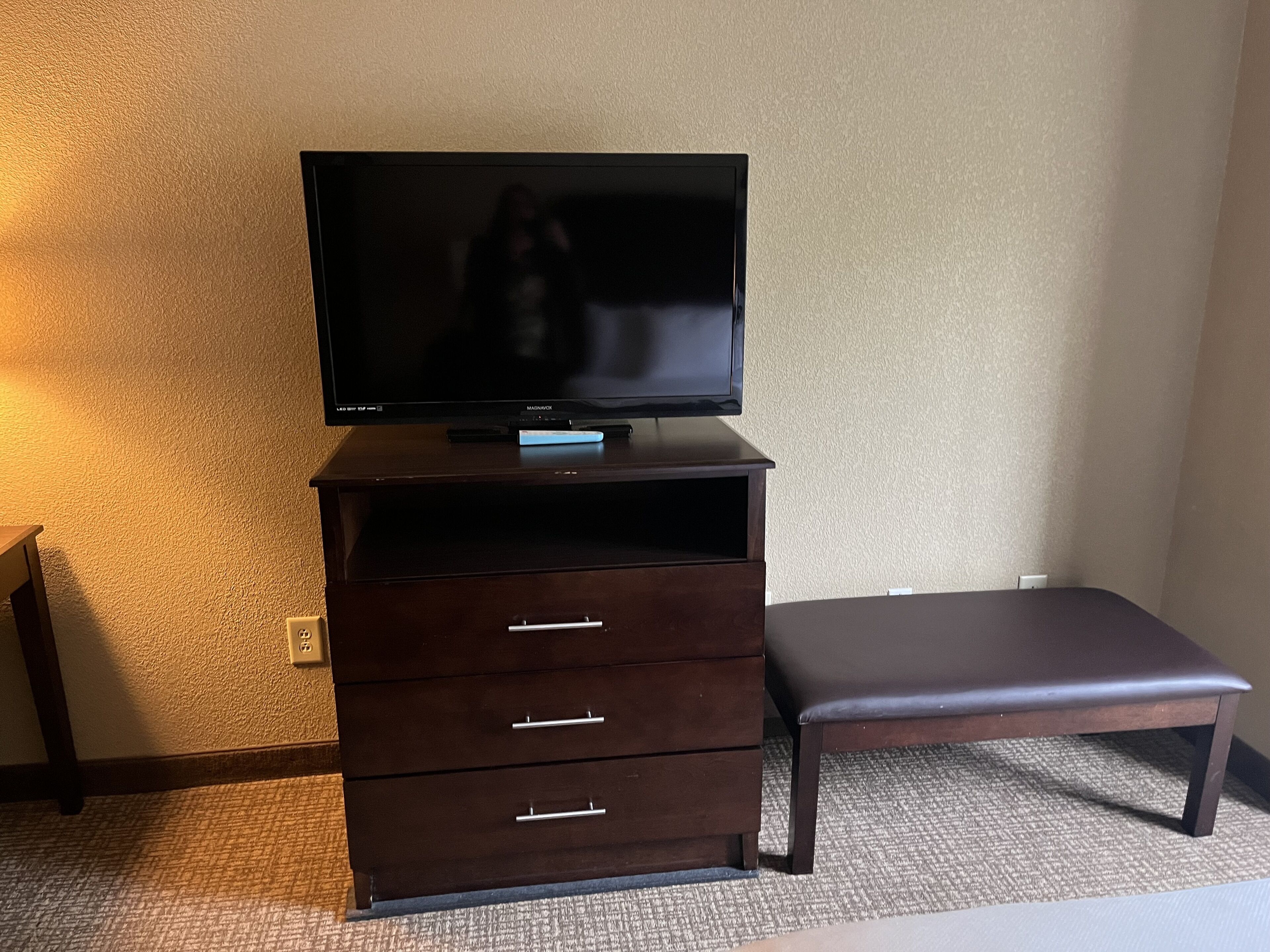 A dark wooden TV stand with a flat-screen TV, a bench, and a small drawer unit against a textured wall.