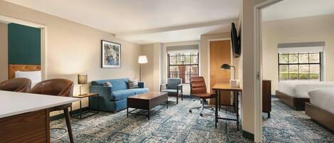A hotel room with a large bed, a desk, a sofa, and a chair. There is a window with a view of trees and a door leading to another room.
