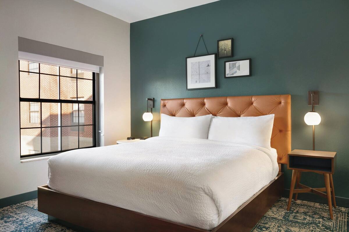 A neatly made bed in a room with a large window, a wall-mounted clock, and a small bedside table with a lamp.