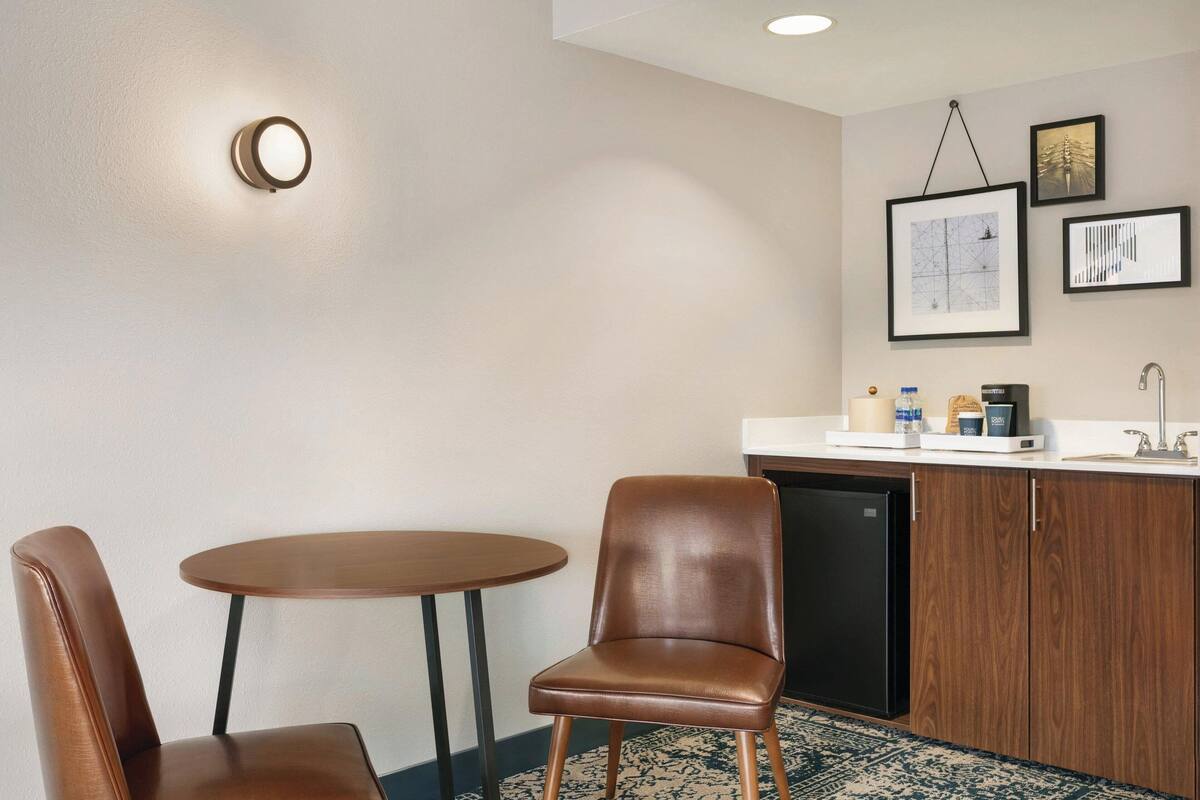 A hotel room with a round table, two brown chairs, a built-in kitchen with a sink, and a refrigerator. There are framed pictures on the wall.