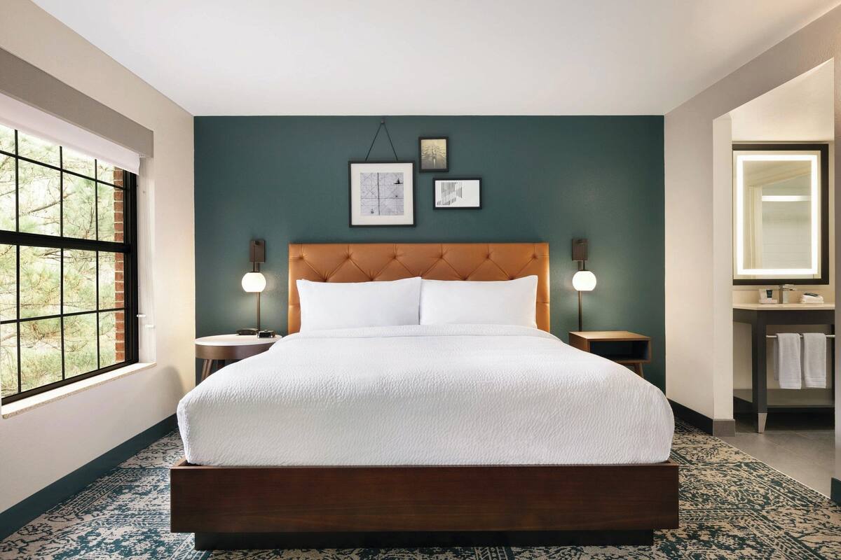 A neatly arranged bedroom with a large bed, a window with a view, and a bathroom with a mirror and sink.
