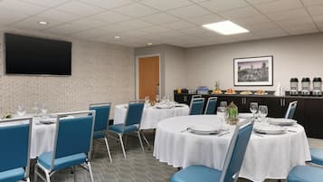 A dining area with tables set for four, blue chairs, white tablecloths, and a flat-screen TV on the wall.