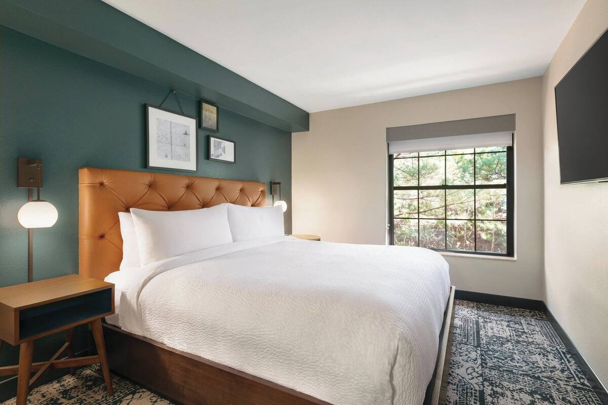 A bedroom with a large bed, a nightstand, a wall-mounted lamp, and a window with a view of trees.