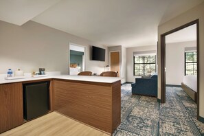 A modern hotel room with a wooden kitchenette, a flat-screen TV, a sofa, and a large window offering a view of trees.