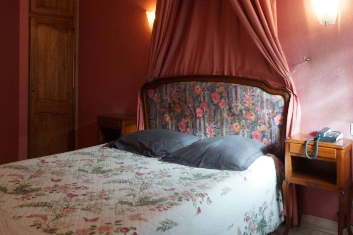 A bedroom with a canopy bed, floral bedding, a wooden wardrobe, and a bedside table with a lamp.