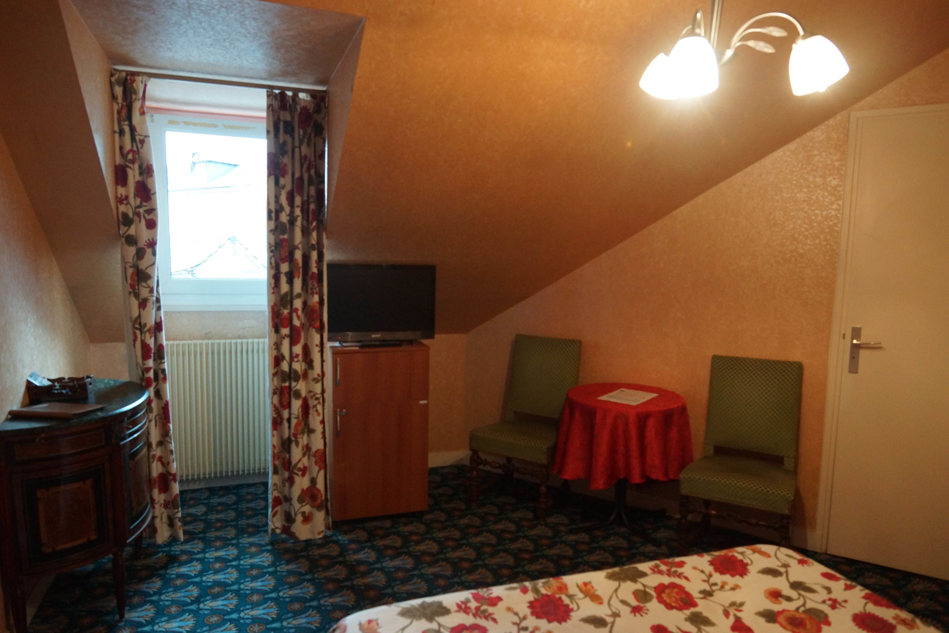 Una habitación de hotel con una cama, un televisor sobre un mueble de madera, dos sillones alrededor de una mesita y una ventana con cortinas.