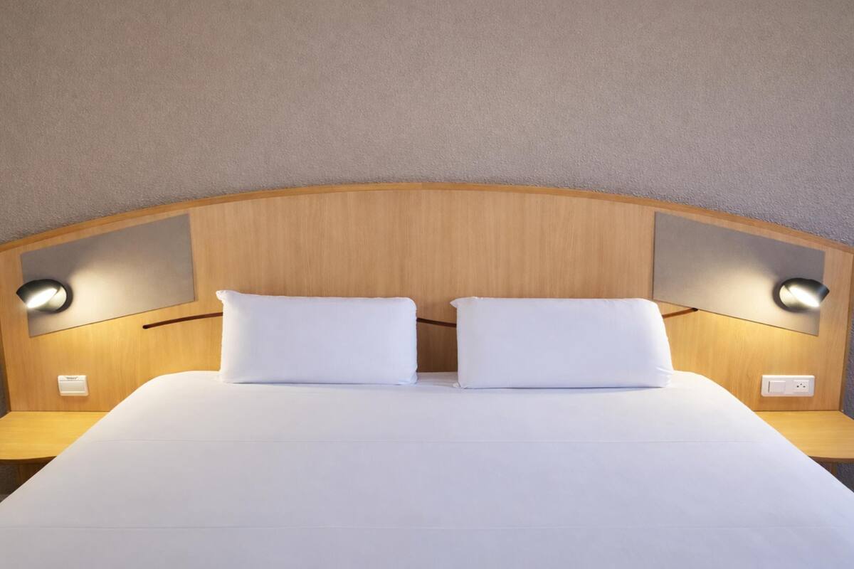 Una cama bien hecha con cabecera de madera, dos mesitas de noche y lámparas de lectura fijadas en la pared.