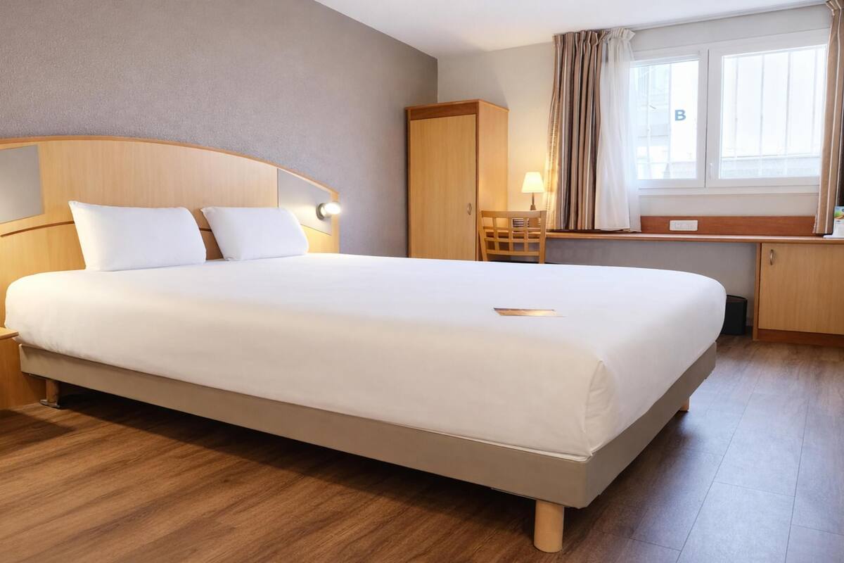 Habitación de hotel con una cama grande, cabecero de madera, mesitas de noche, un escritorio y una ventana con cortinas.