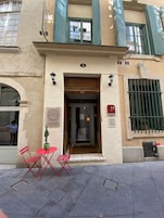 Entrada de hotel con puerta de madera, un pequeño espacio al aire libre con sillas rojas y una planta en maceta.