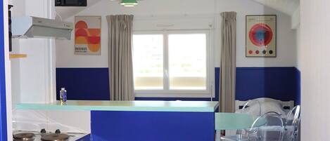 A compact kitchen with a blue accent wall, a small dining area with clear chairs, and a window with curtains.